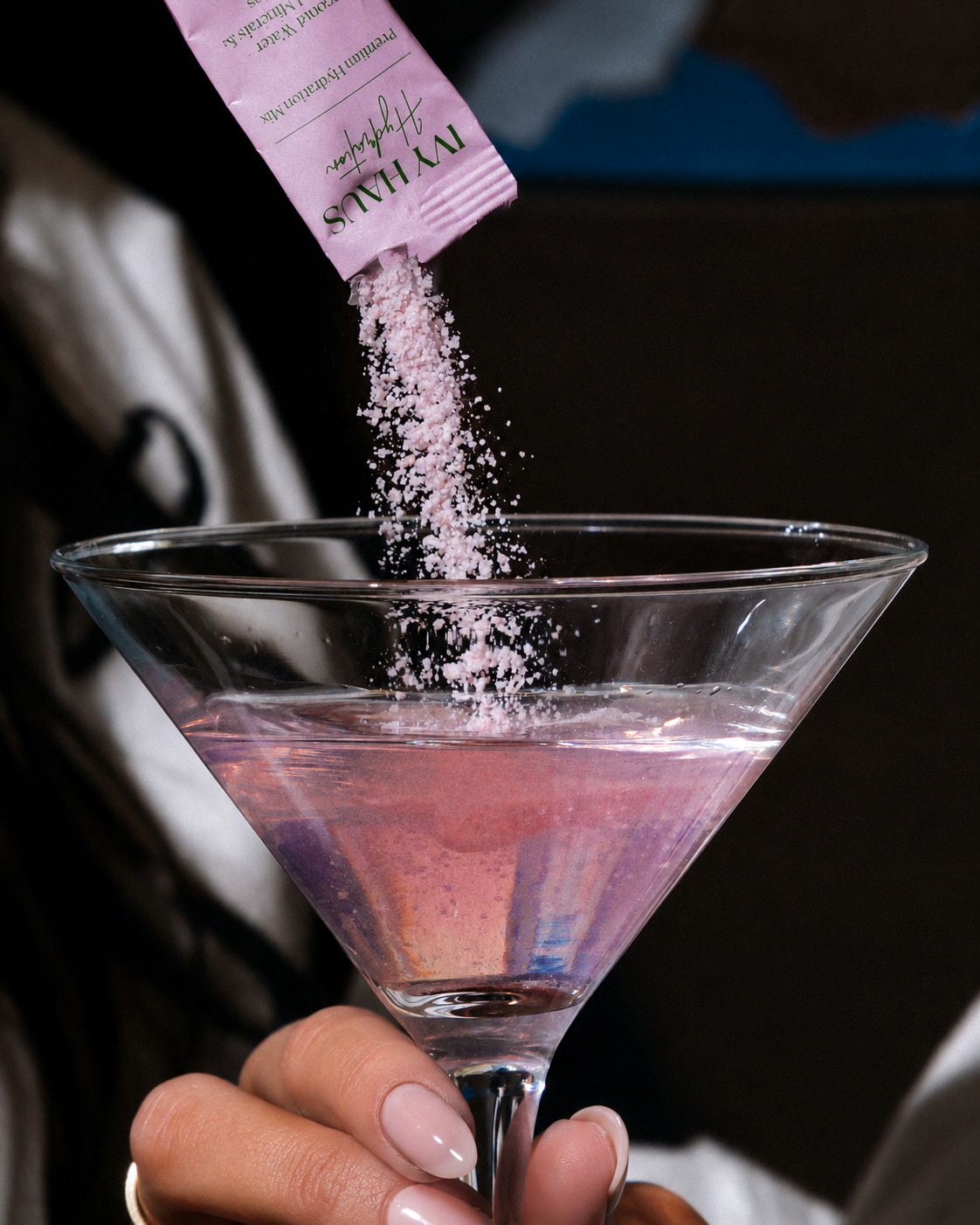 A hand holding a martini glass filled with pink liquid, with a pink packet pouring light pink powder into the drink. The packet shows the brand name 'ivy haus.
