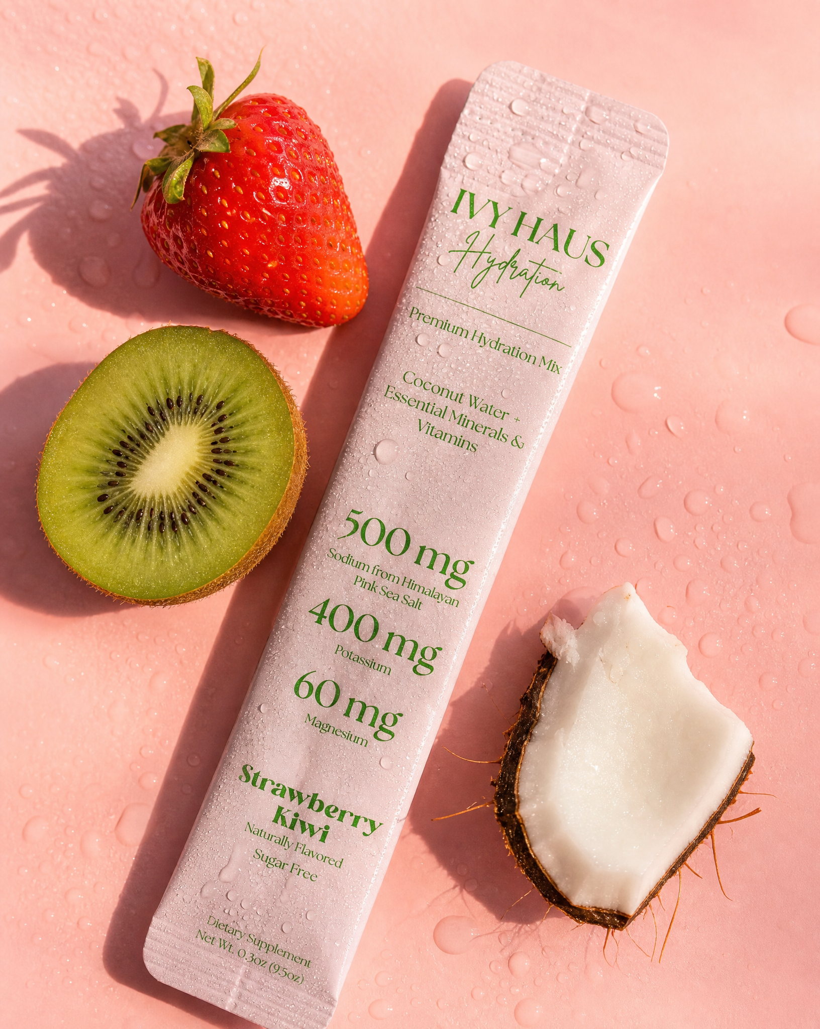A single IVYHAUS Hydration Strawberry Kiwi powder sachet lies on a pink surface with water droplets, styled with a whole strawberry, a sliced kiwi, and a coconut half nearby.