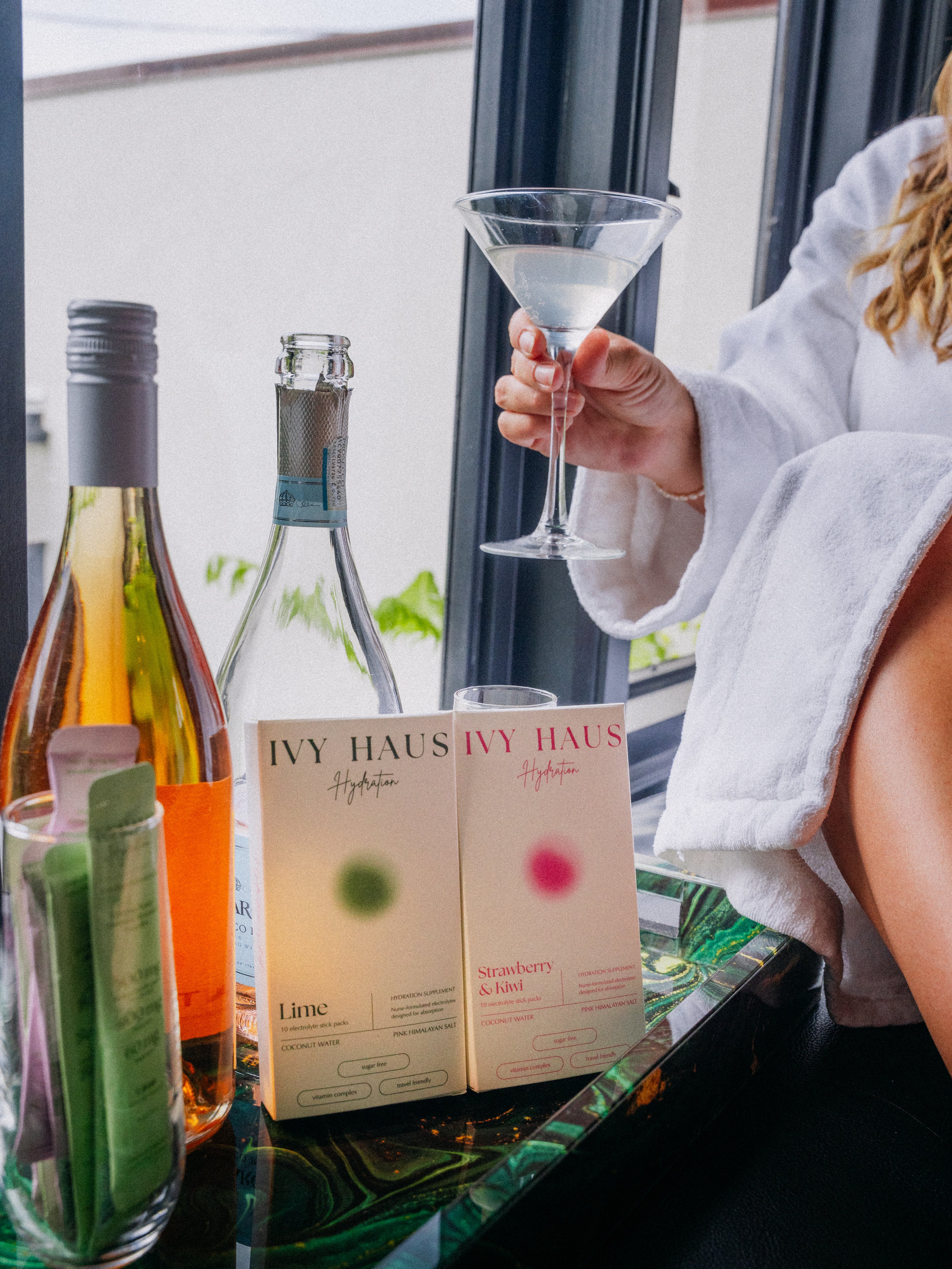Two IVY HAUS Hydration drink boxes on a glass table next to wine bottles, with a person in a white robe holding a martini glass by a window.