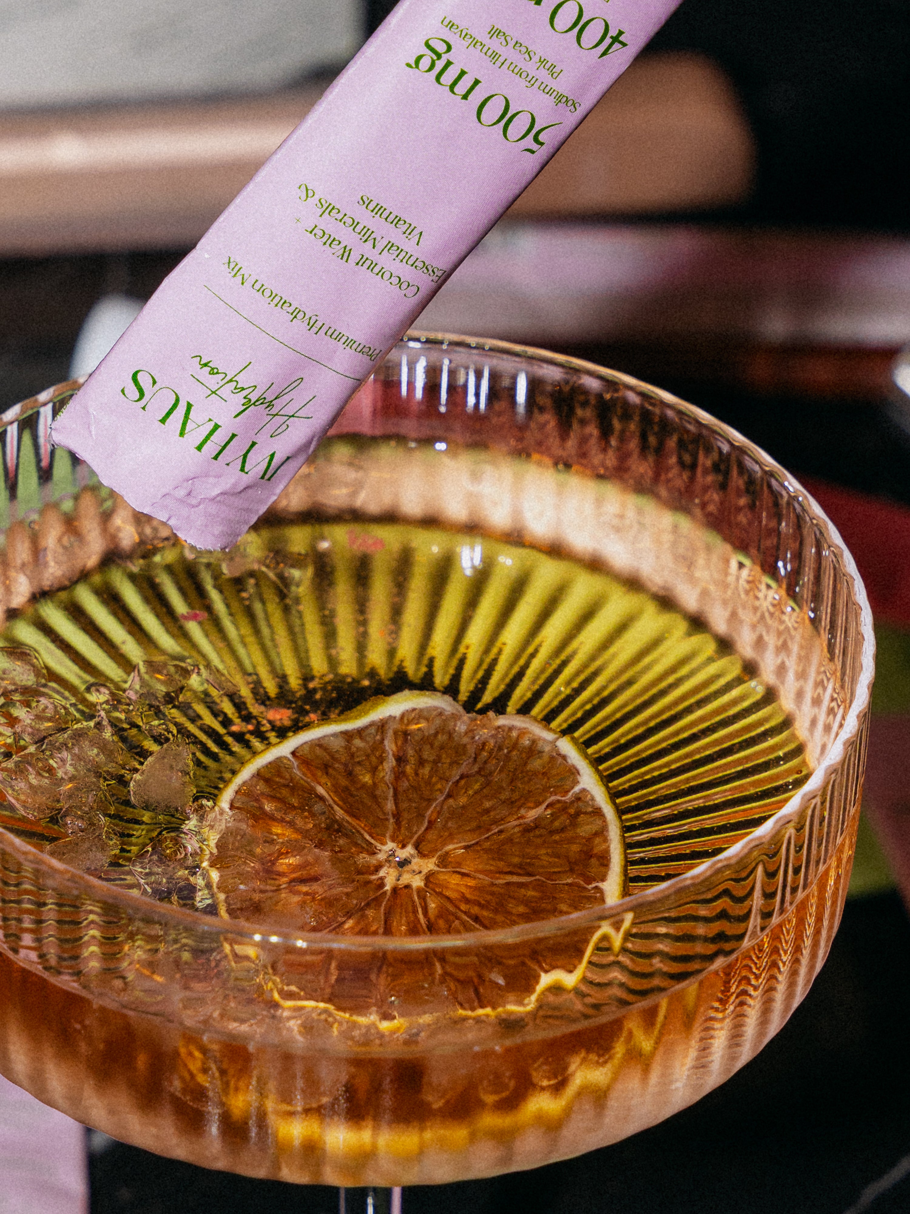 A close-up of a cocktail in a ribbed coupe glass with a lemon slice floating on the surface, with a pink sachet labeled 'ivy haus' being poured into the drink.