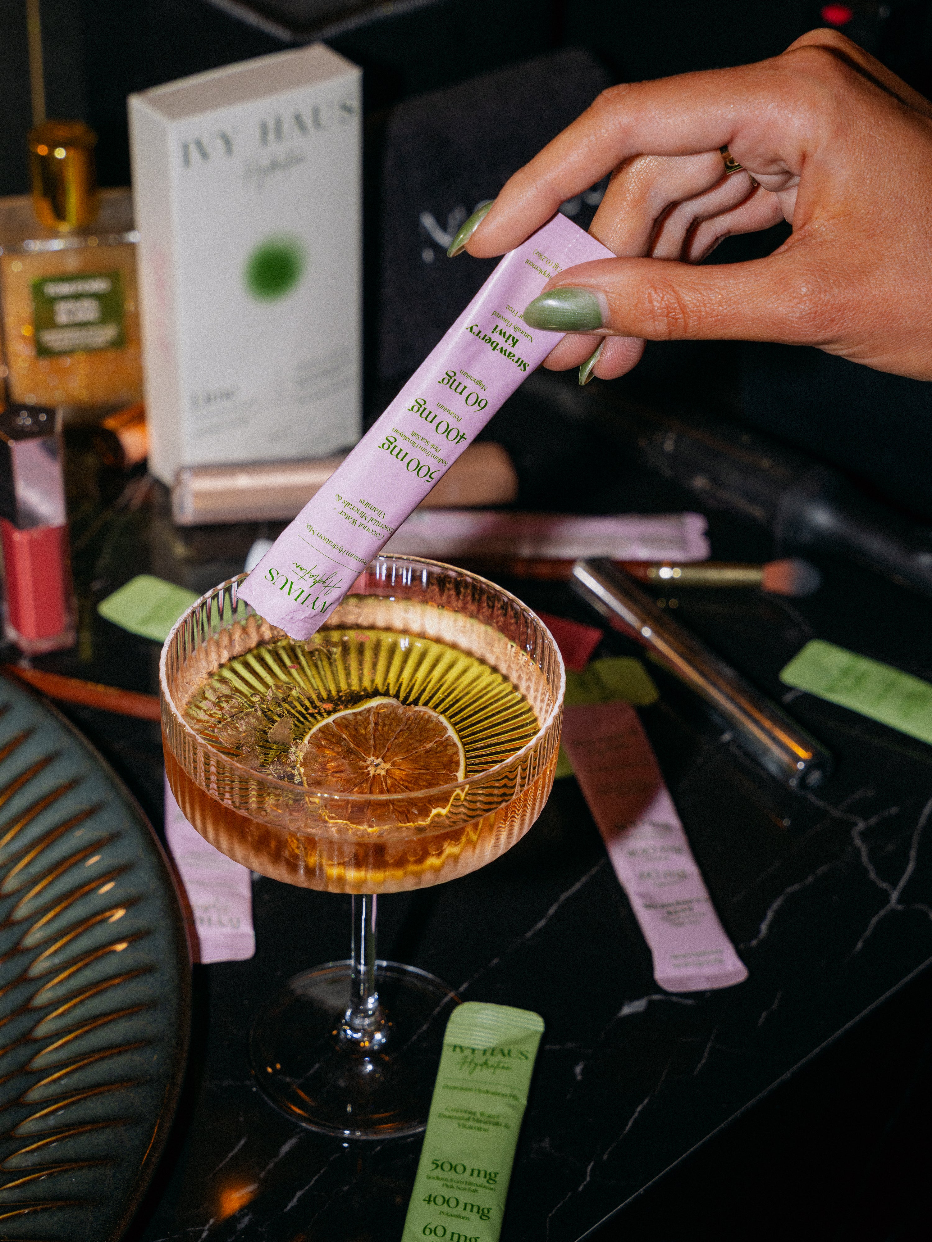 A hand with green nails pouring a pink ivy haus satchet into a cocktail in a coupe glass, with other beauty products and tubes on a black marble surface.