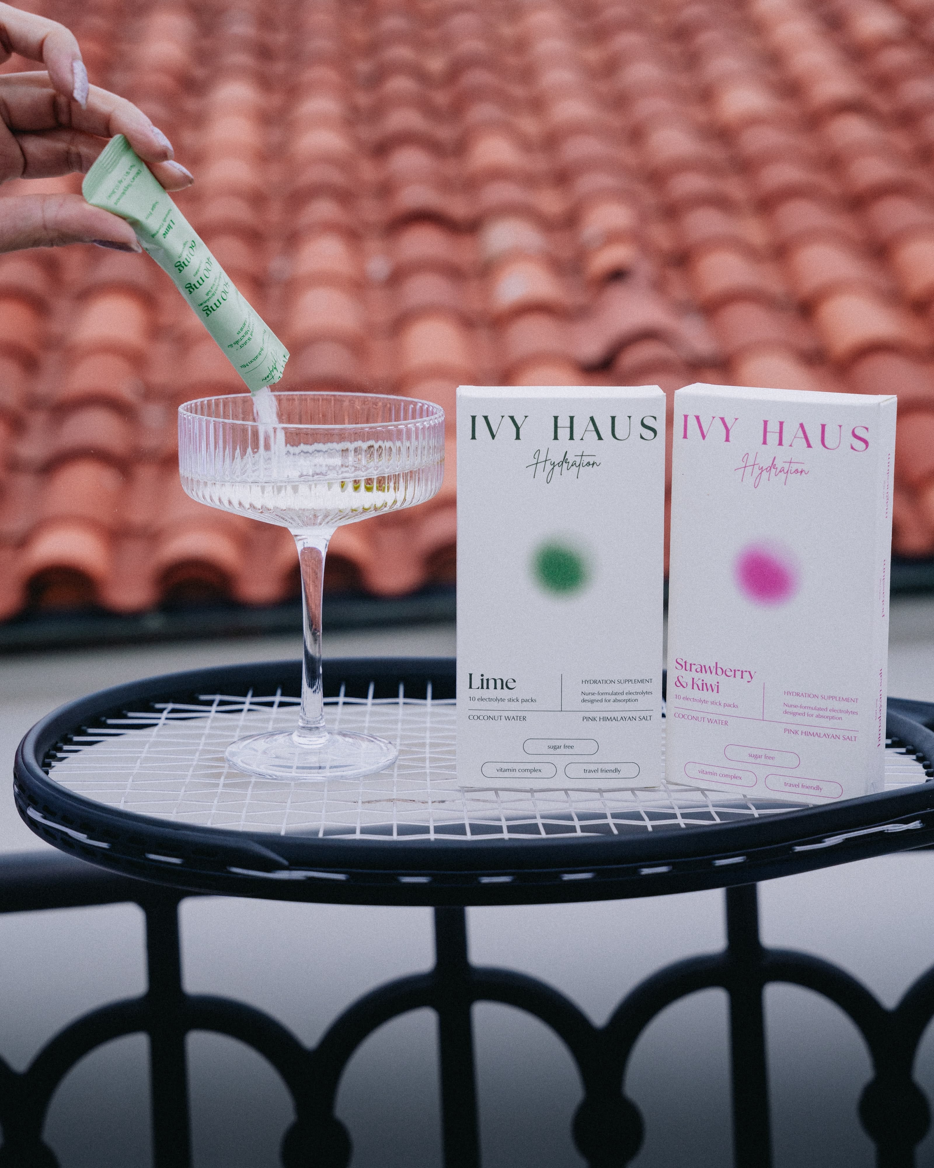 A hand pours a green tube of IVY HAUS Hydration packet into a coupe glass, with two product boxes (Lime and Strawberry & Blue) standing on a tennis raquet in front of a terracotta tiled roof background